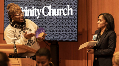 Woman on the left stands behind a podium, holding an award, while speaking to the woman on the right who will soon receive the award..