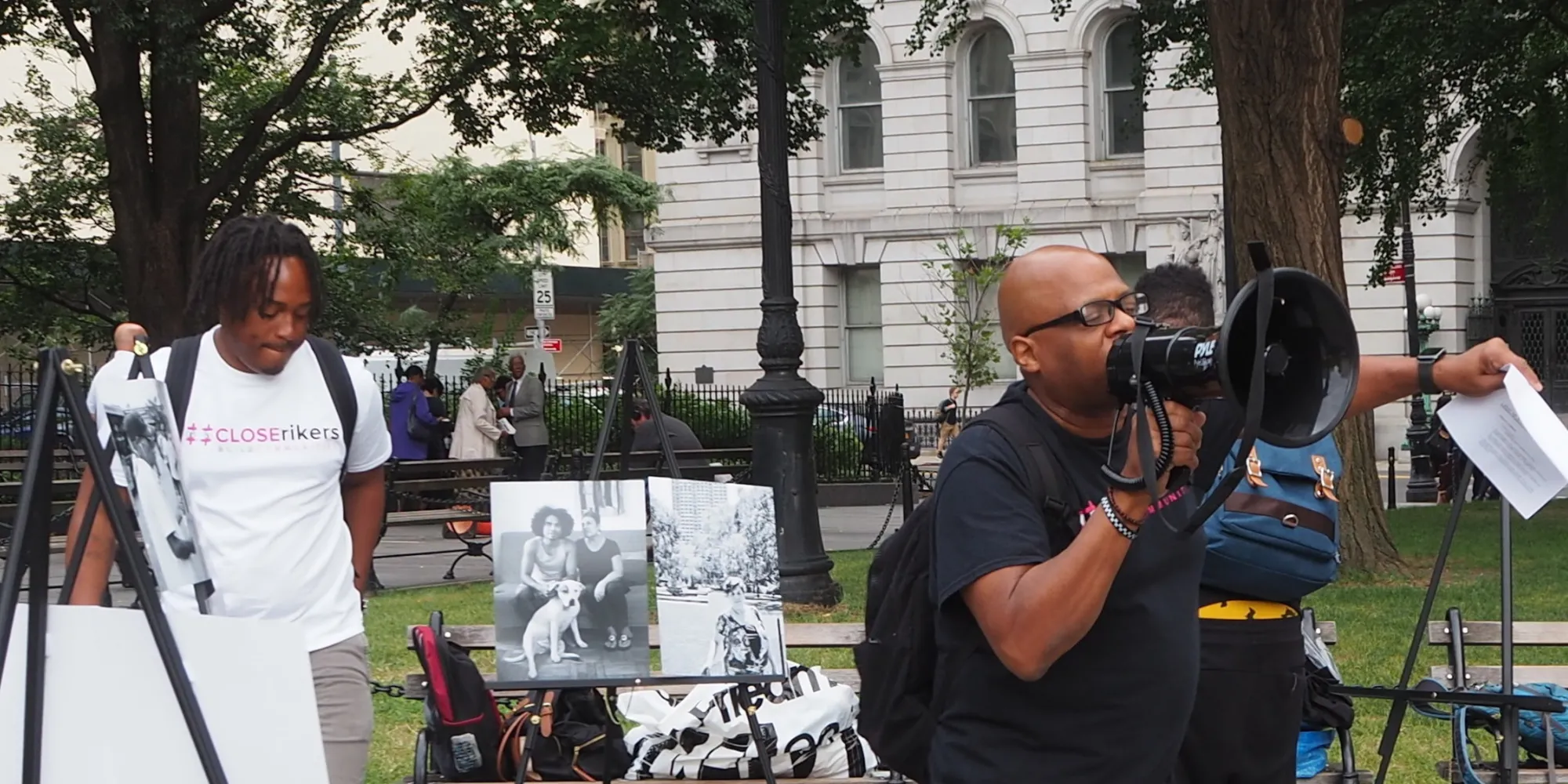 Close Rikers protest in front of City Hall. A man with locs looks down in the background, while another bald man in glasses holds a megaphone.